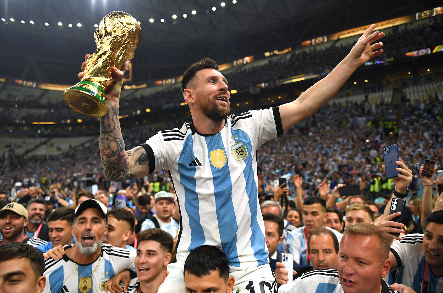 Lionel Messi in a striped light blue and white jersey holding the World Cup Trophy being lifted on the shoulders of his teammates. 